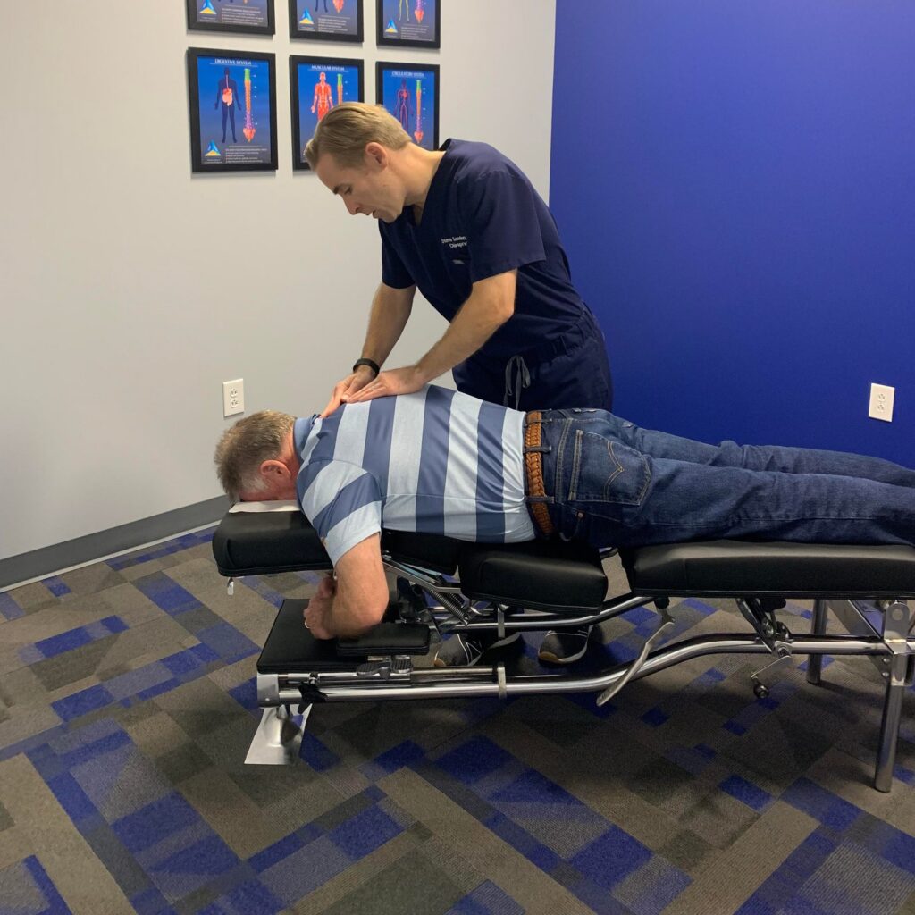 Chiropractor adjusting patient's lower back on treatment table in clinical room with anatomy charts